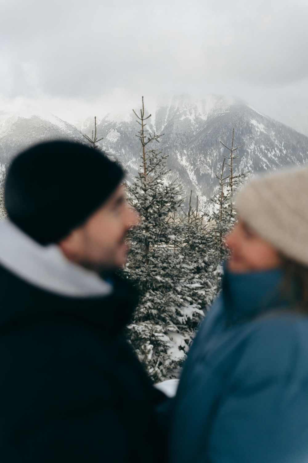 Couple in winter hats gazing at each other with snow-covered pine trees and misty mountains in the background.