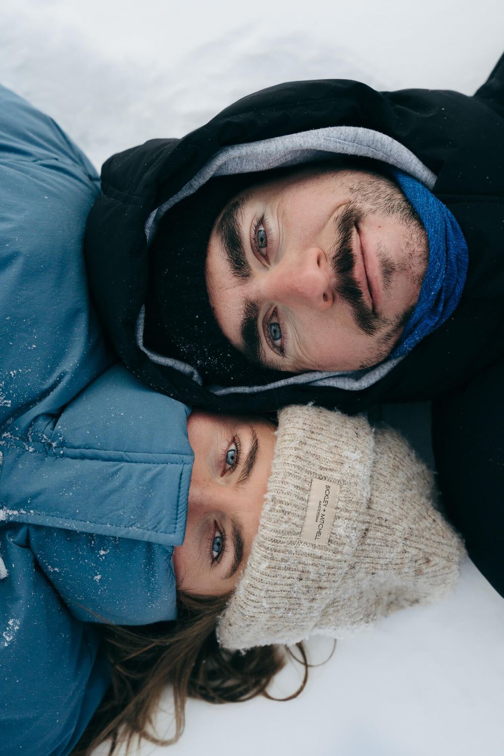 Couple lying in snow wearing winter hats and jackets, smiling together on a snowy day.
