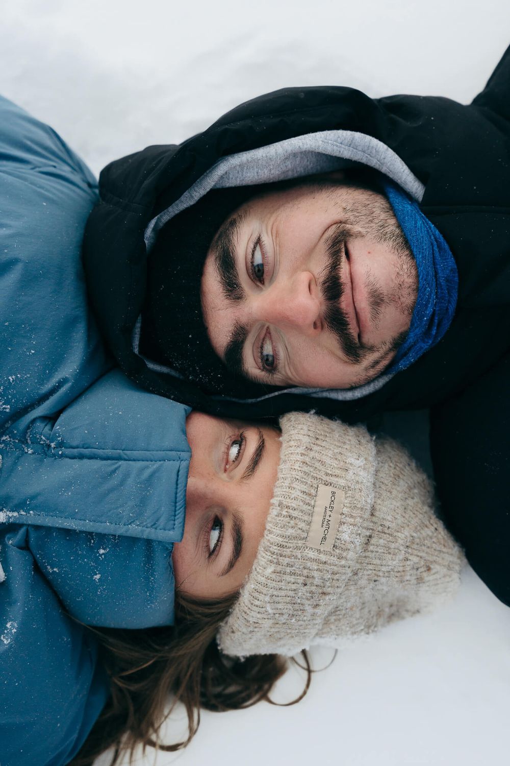 Couple lying in snow wearing winter jackets and hats, smiling together outdoors in a snowy landscape.