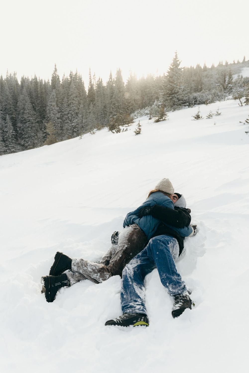 Couple embracing and playing in snow on a winter mountain slope surrounded by pine trees.