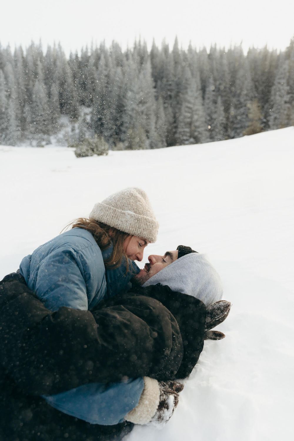 Couple sharing a romantic moment lying in snow, surrounded by snow-covered pine trees in a winter landscape.