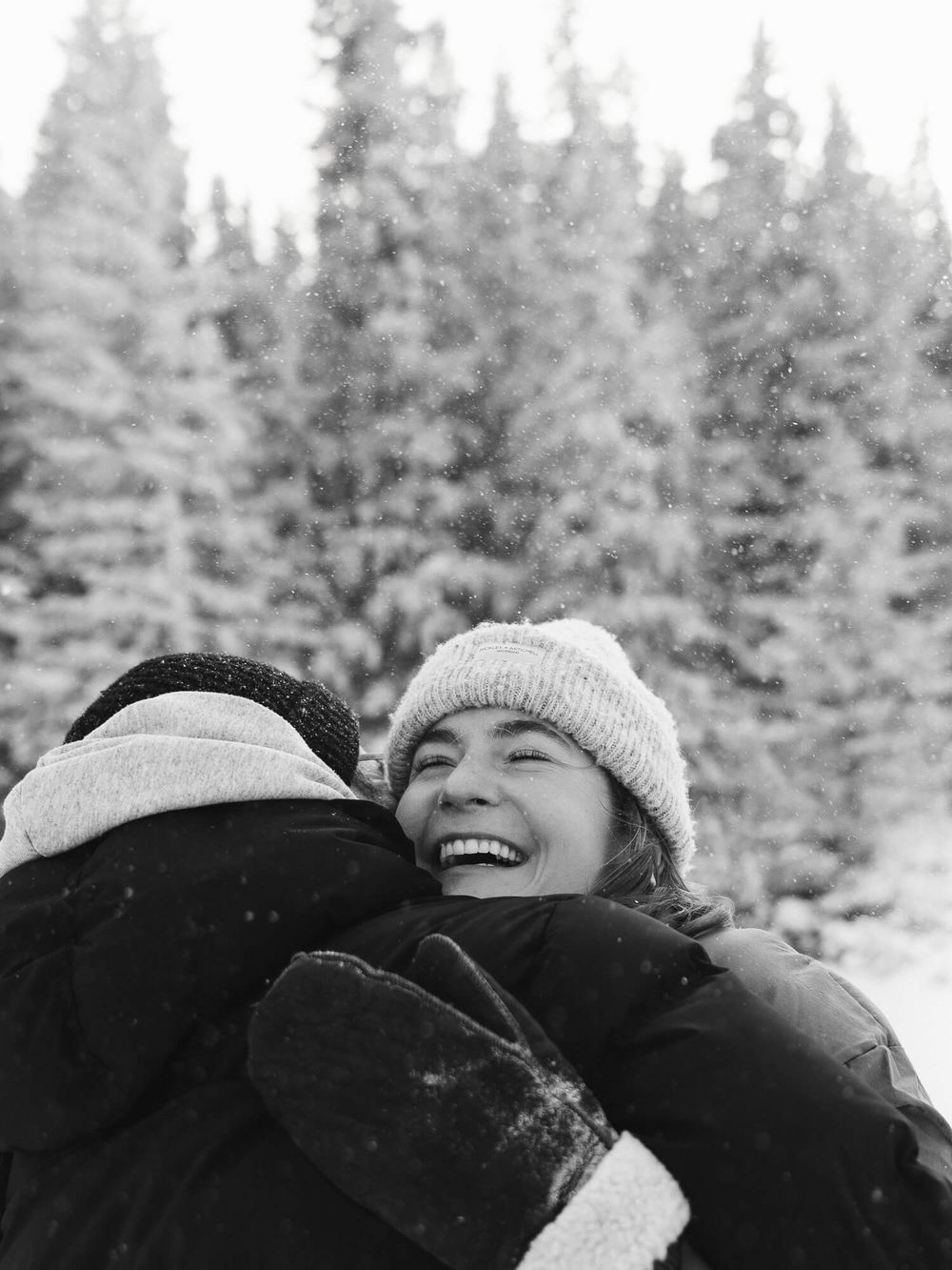 Two people hugging and laughing in a snowy winter landscape with snow-covered evergreen trees in the background.
