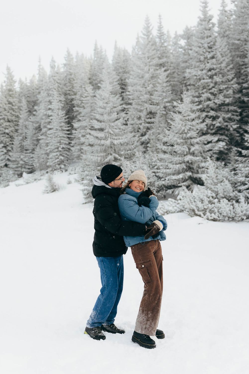 Couple embracing and kissing in snowy winter landscape with snow-covered evergreen trees in background.