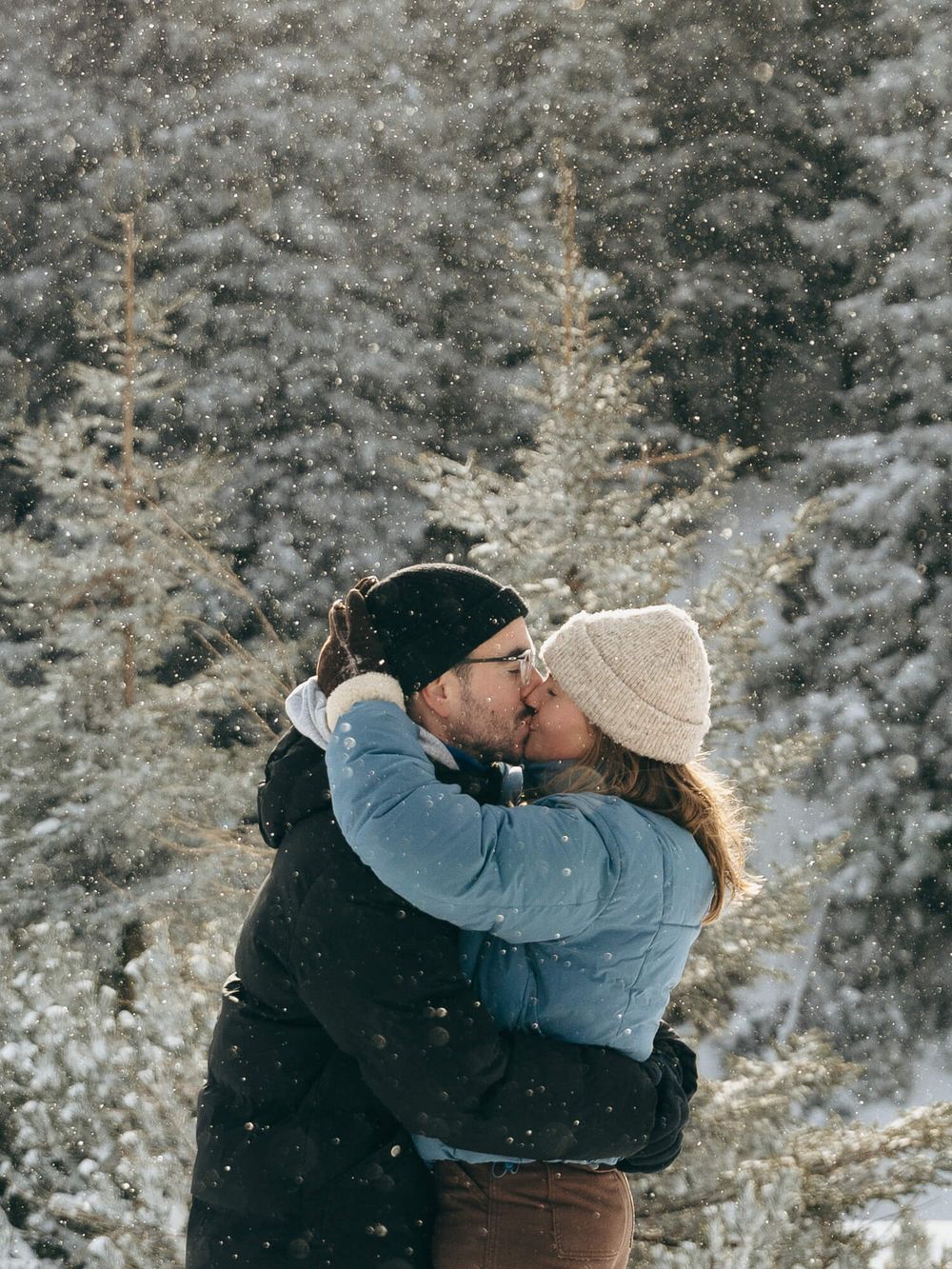 Couple sharing a romantic kiss in falling snow with snow-covered pine trees in the background.