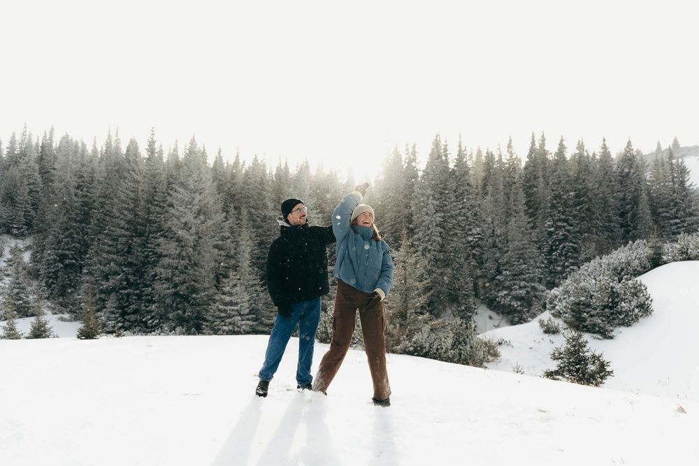 Two people standing in a snowy winter landscape with snow-covered pine trees in the background.