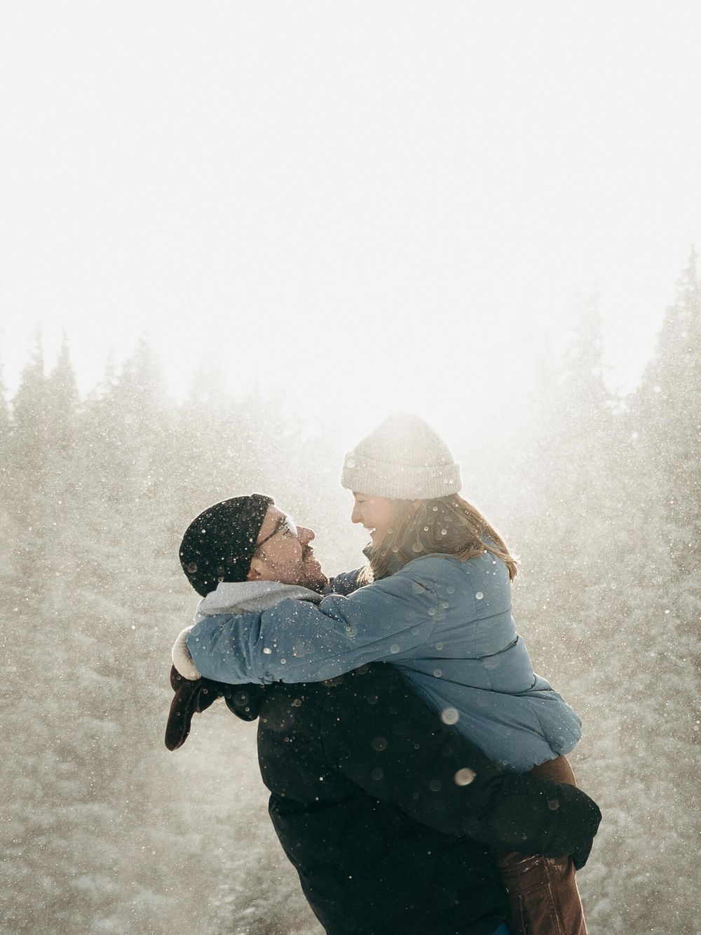 Couple embracing and laughing in a snowy winter wonderland surrounded by snow-covered evergreen trees.