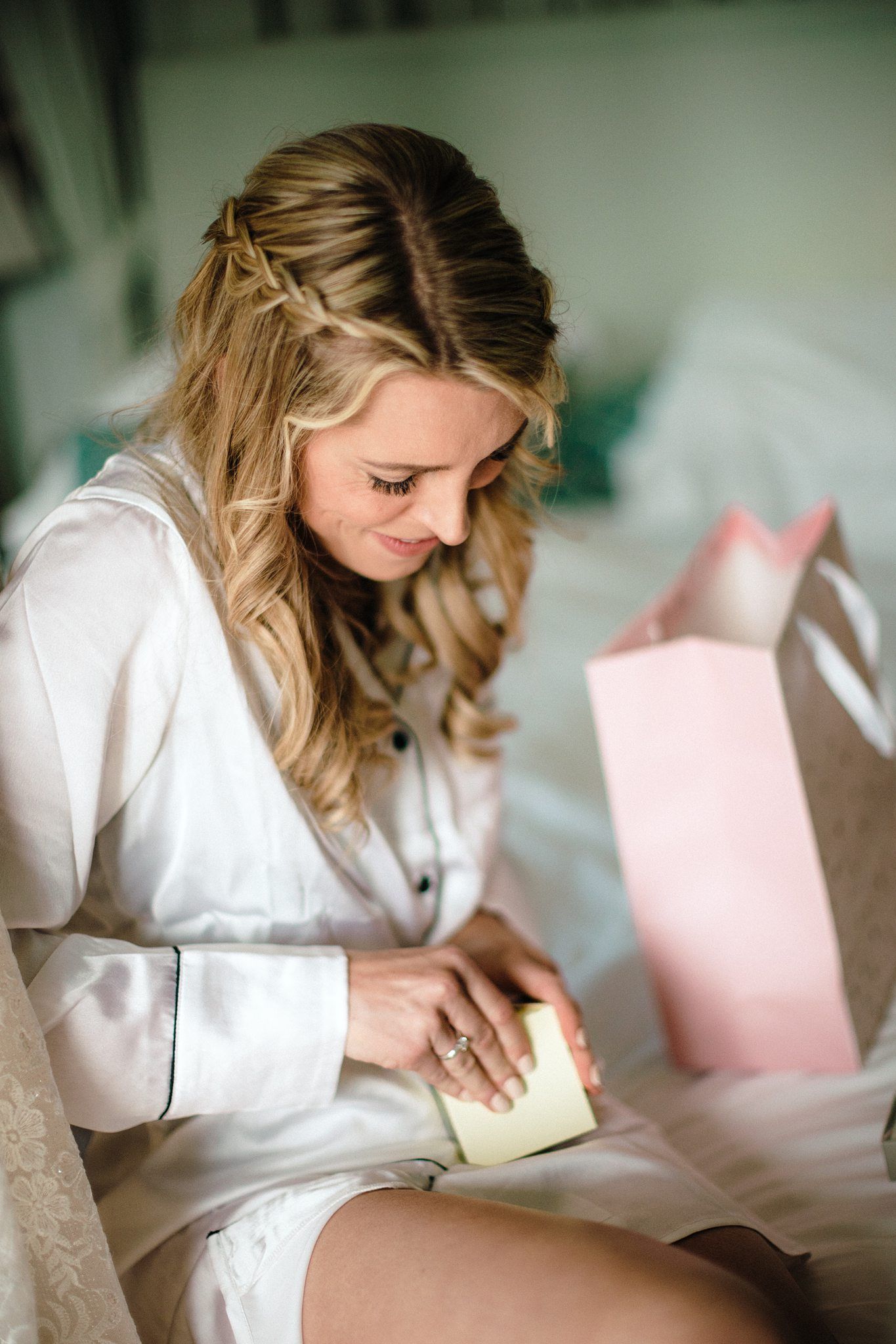 Bride in white satin robe reading a card from a gift bag while sitting on a bed before her wedding at The Prince George Hotel, Halifax.