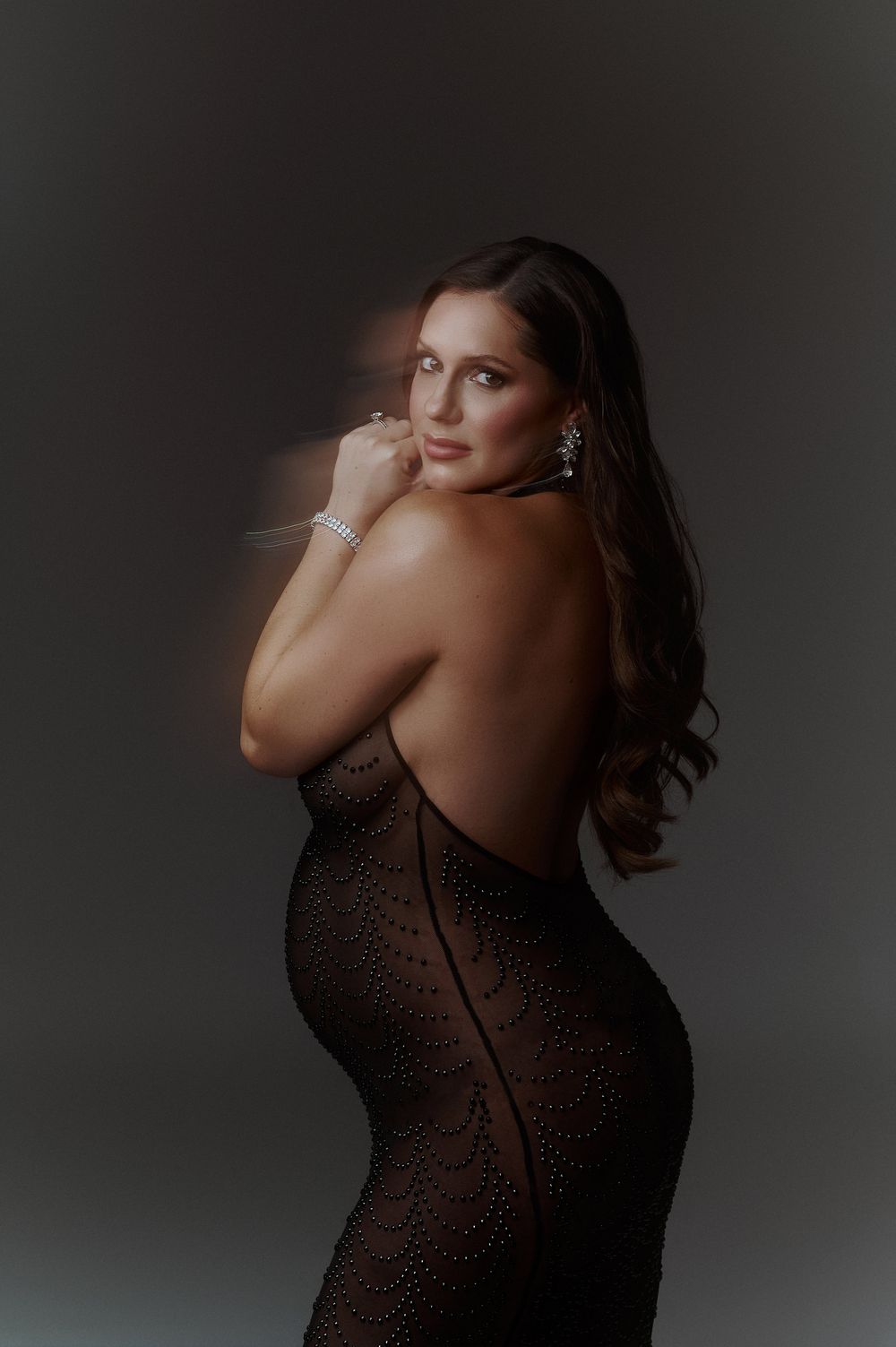 Elegant pregnant woman in backless black lace gown with diamond jewelry in a studio portrait.