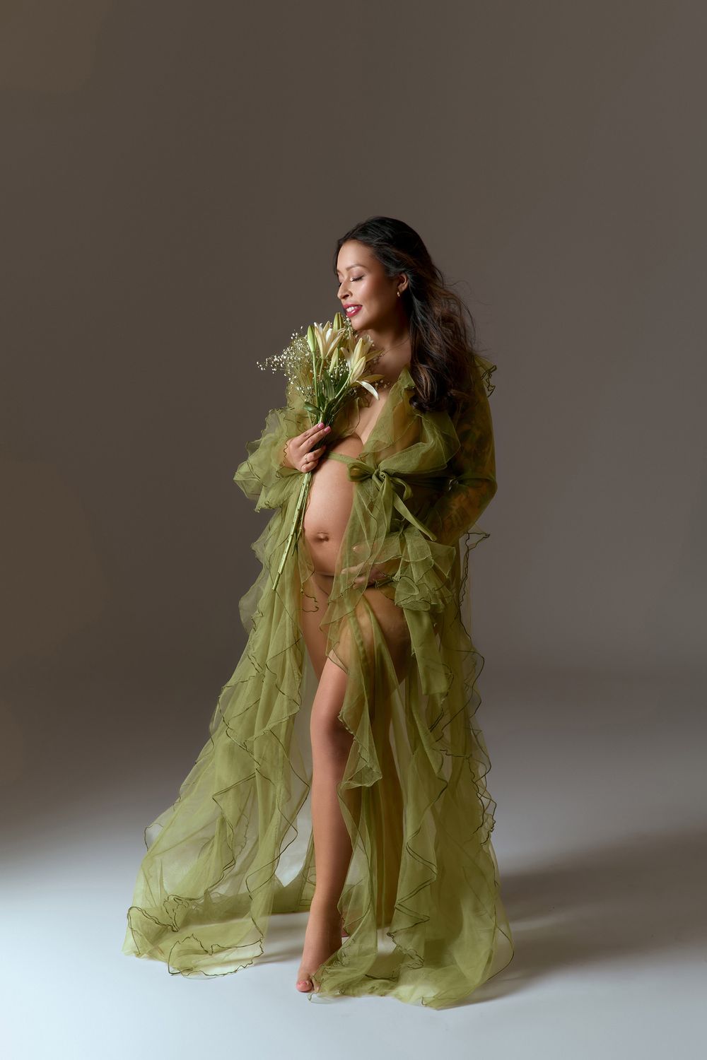 Pregnant woman in sheer green ruffled robe holding wildflowers in a studio maternity photo.