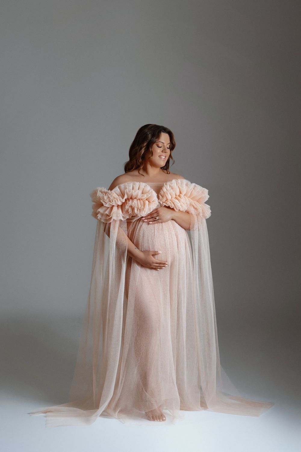 Pregnant woman in off-shoulder blush pink ruffle tulle gown cradling belly in studio portrait.