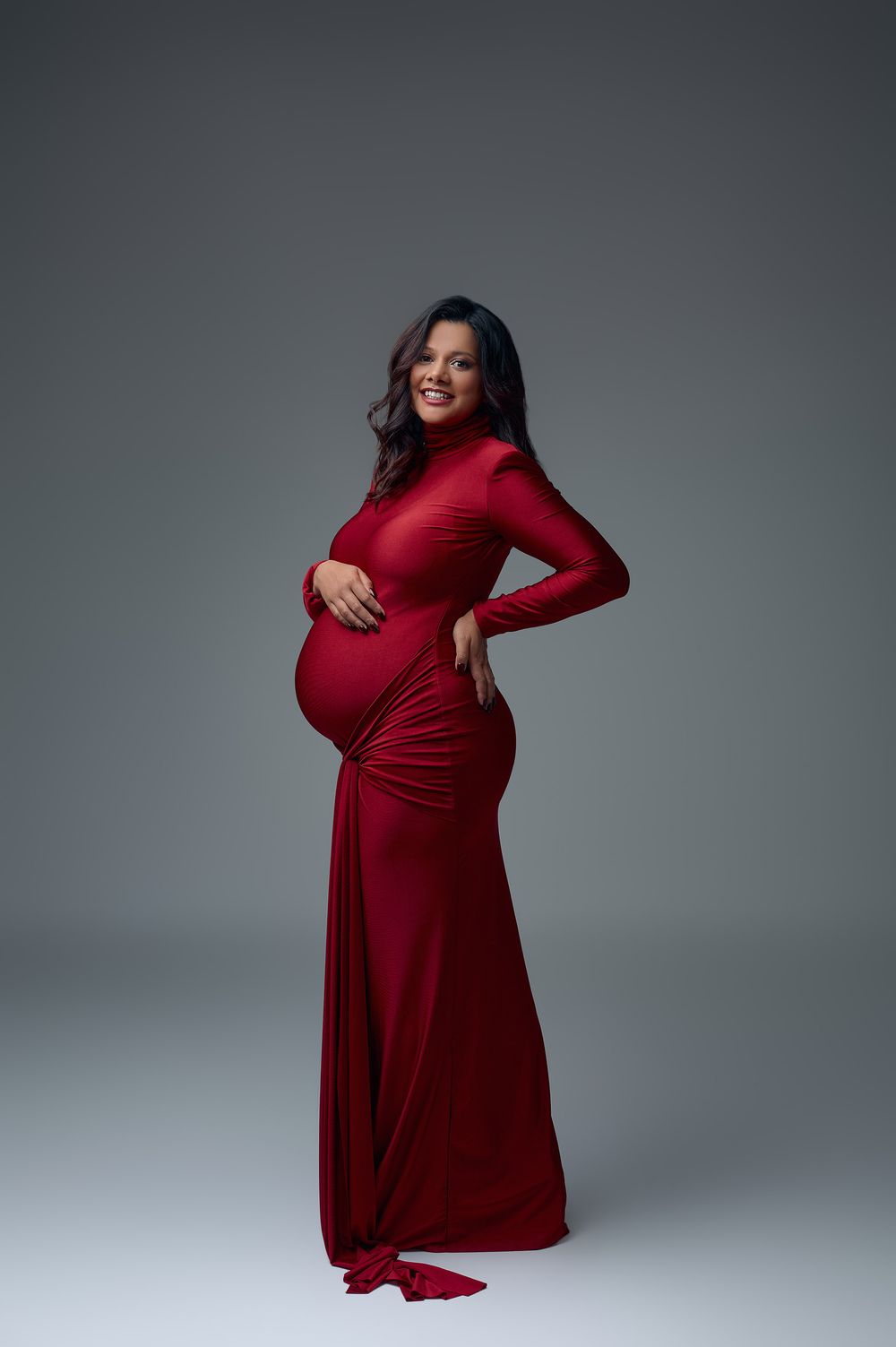 Smiling pregnant woman in elegant red long-sleeve gown posing in studio, cradling baby bump.