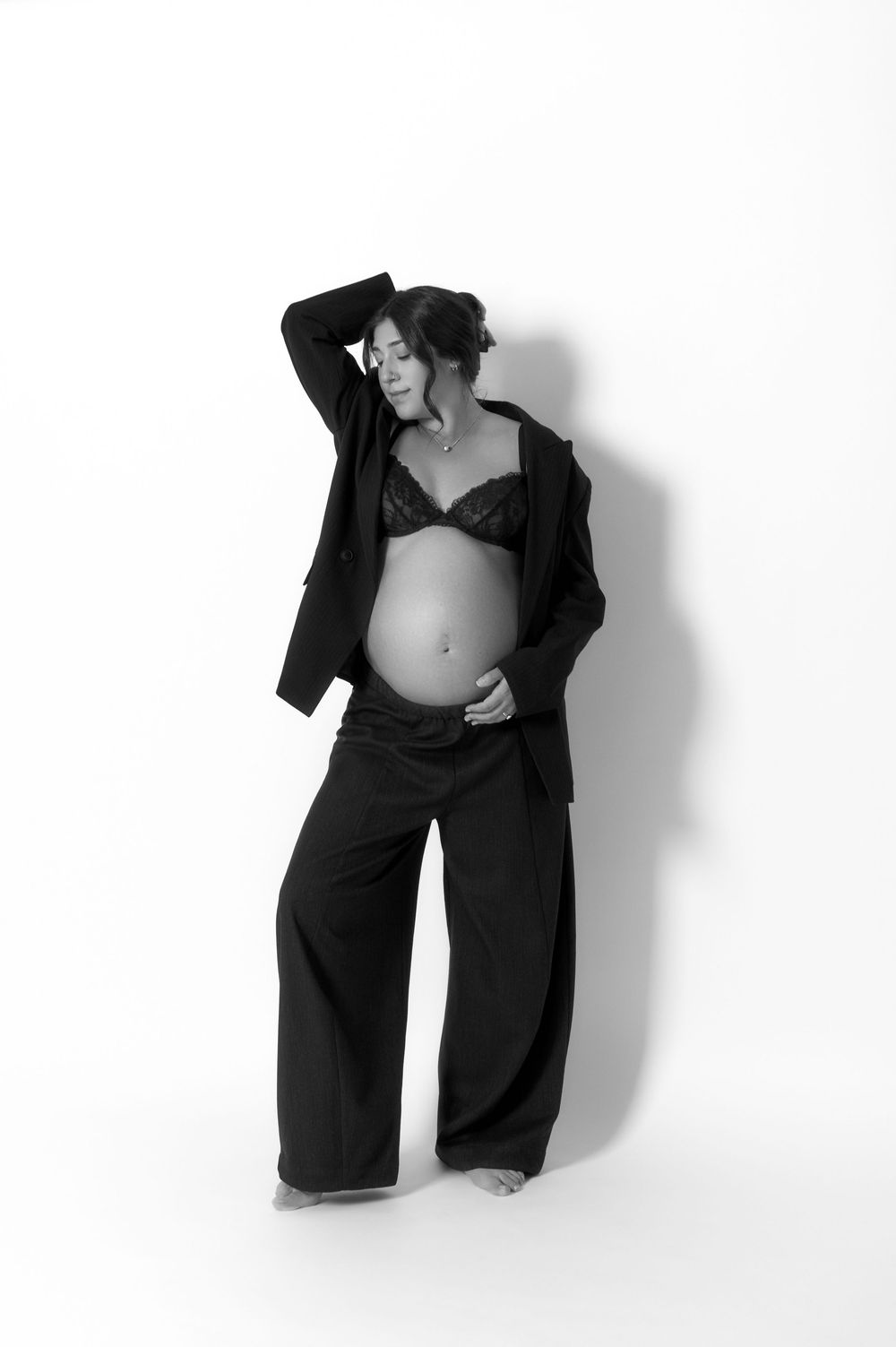 Black and white maternity photo of pregnant woman in black suit jacket, bra, and wide-leg pants.