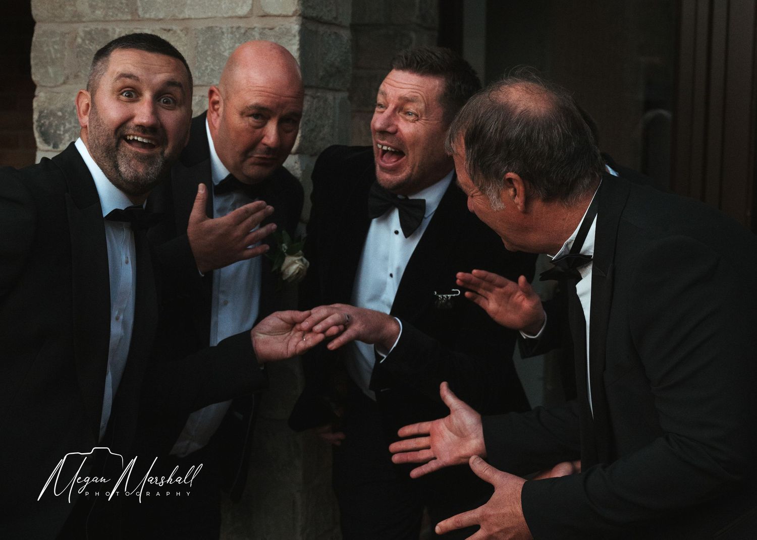 Five men in formal black tuxedos laughing and gesturing together in a black and white wedding photo.