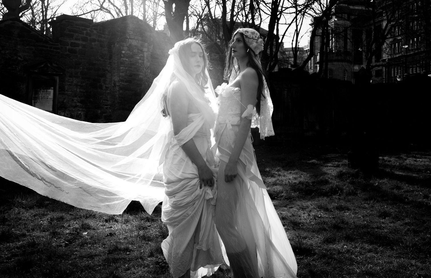Two brides in white wedding gowns and veils pose outdoors in a black and white photo, veil flowing in the wind.