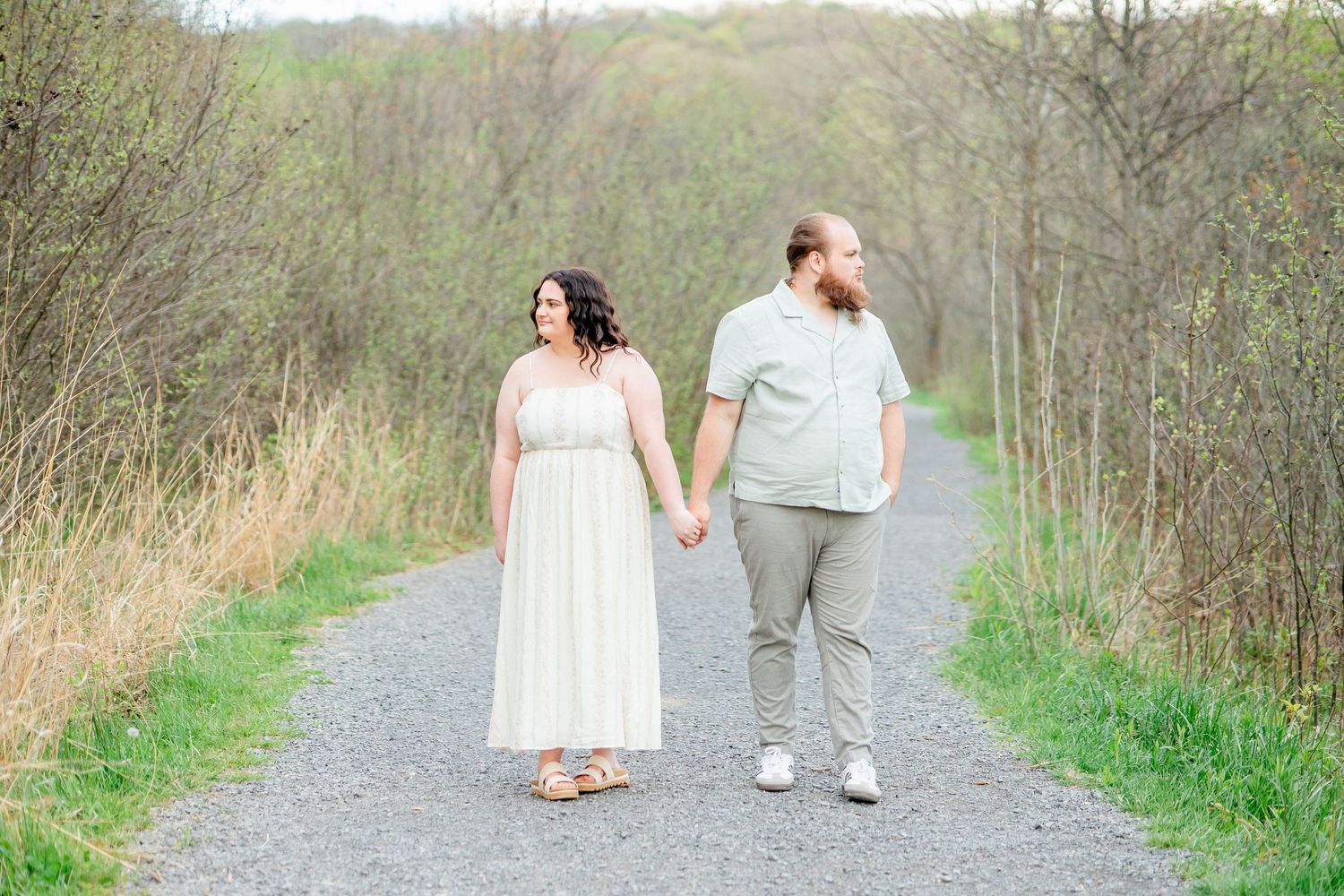 Couple holding hands walking on a gravel path surrounded by lush greenery, woman in white dress, man in sage shirt.