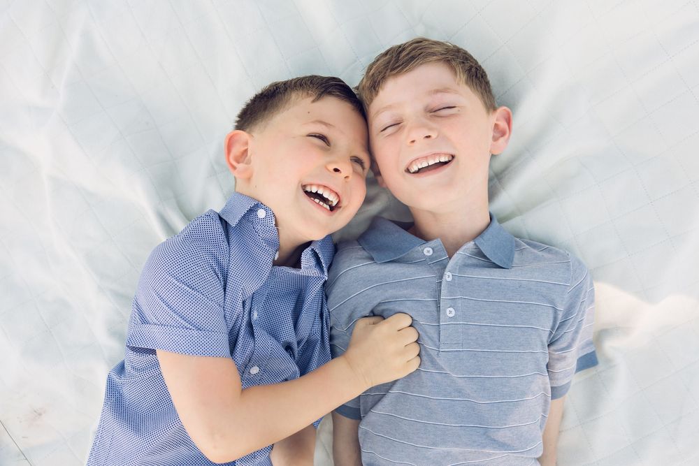 Two young brothers laughing joyfully together while lying on a white blanket in a fun candid moment.