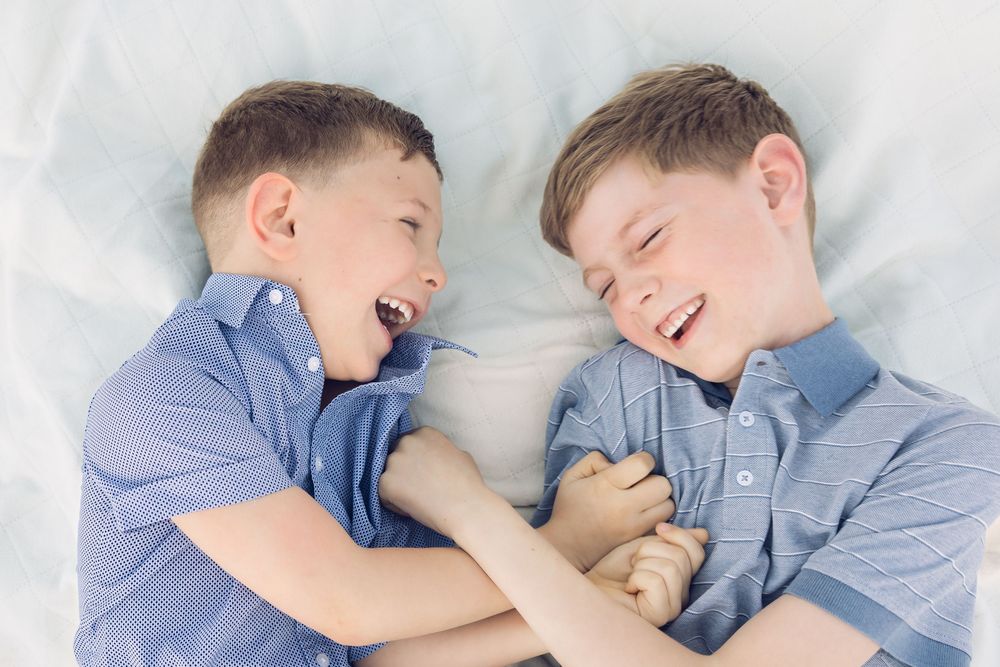 Two young boys in blue shirts laughing and playing together while lying on a soft white blanket.