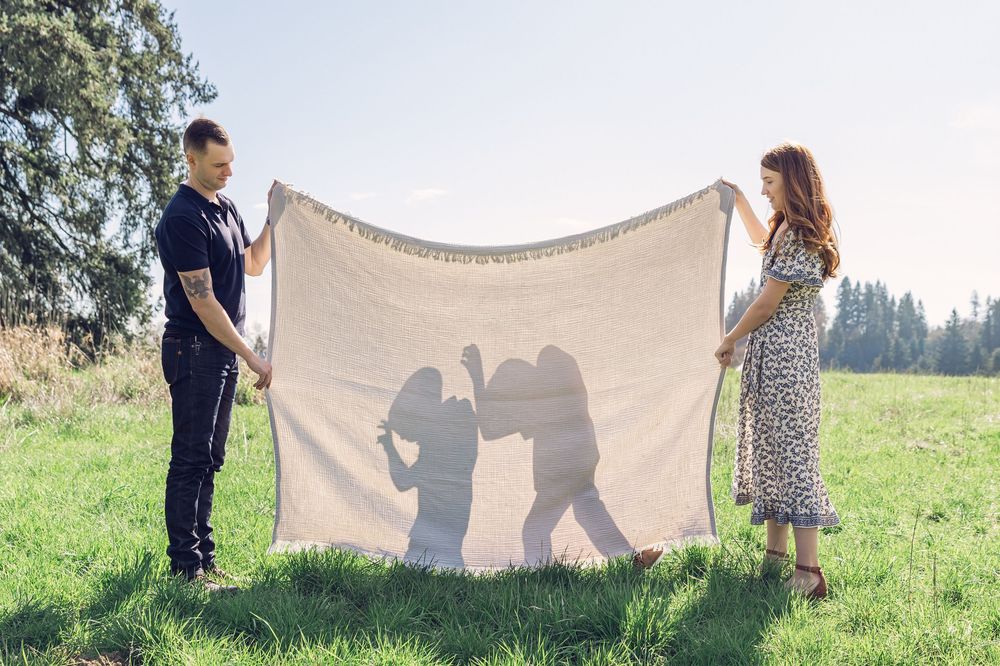 Couple holding up a large blanket outdoors in a green field with childrens silhouettes projected on it.