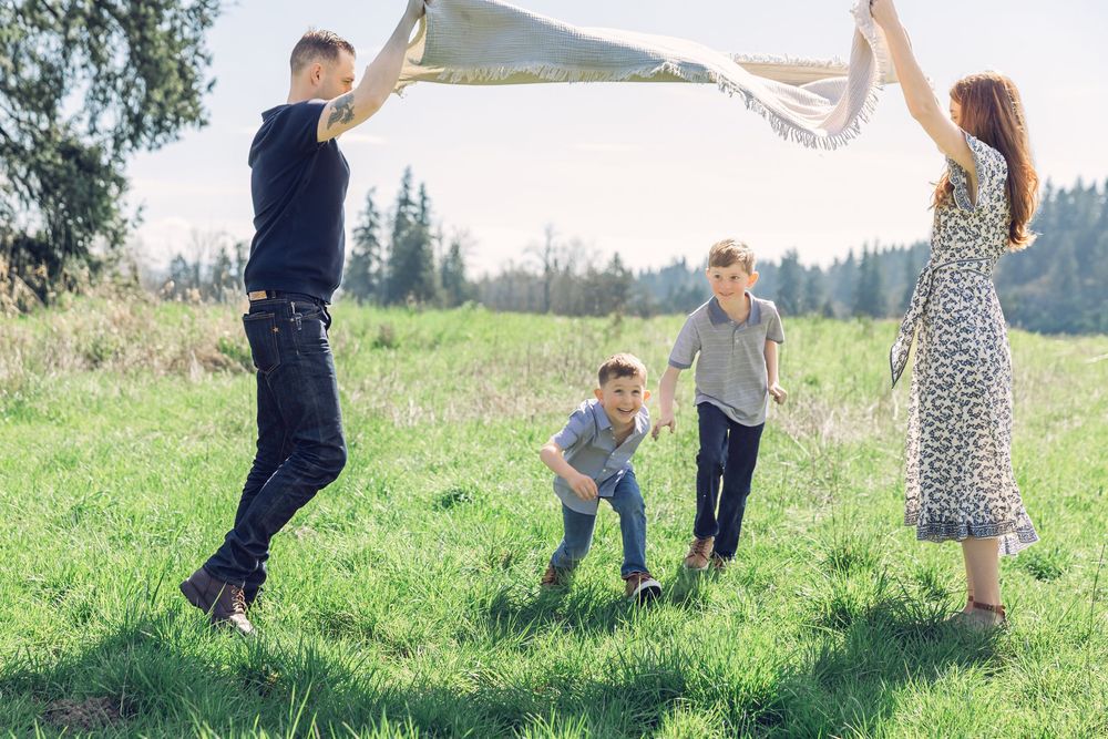 Family of four playing outdoors in a sunny green meadow, waving a blanket over two running boys.