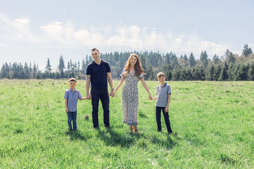 Family of four holding hands while standing together in a wide sunny green meadow with pine trees.