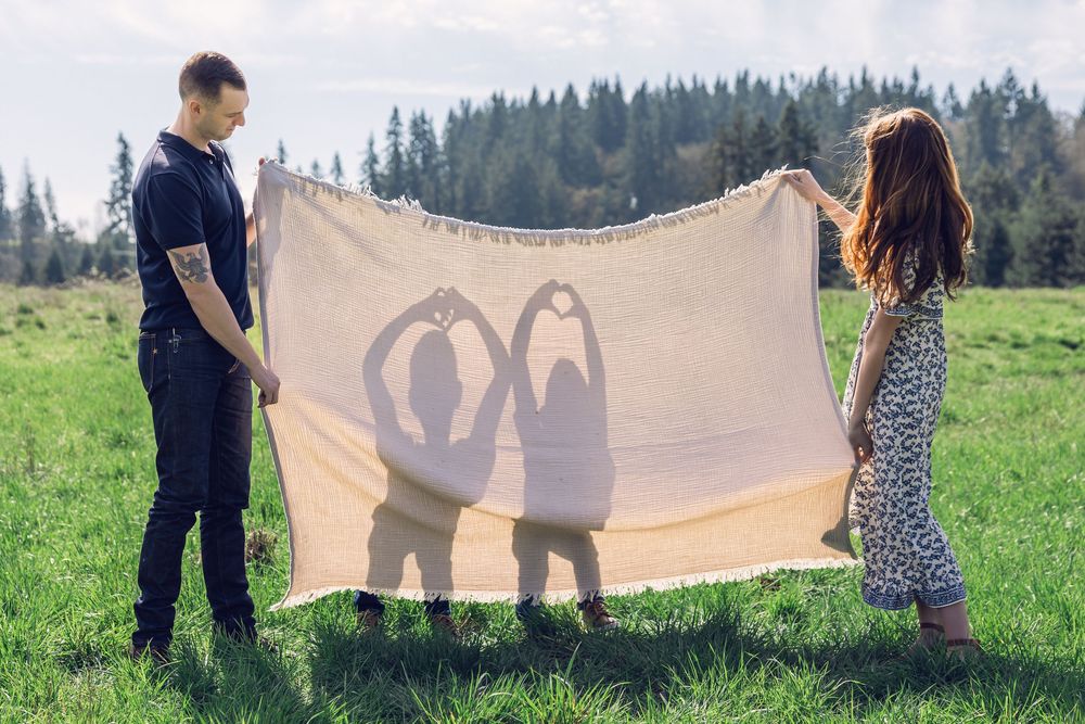 Couple holds blanket showing silhouettes of two children making heart shapes in a green mountain meadow.