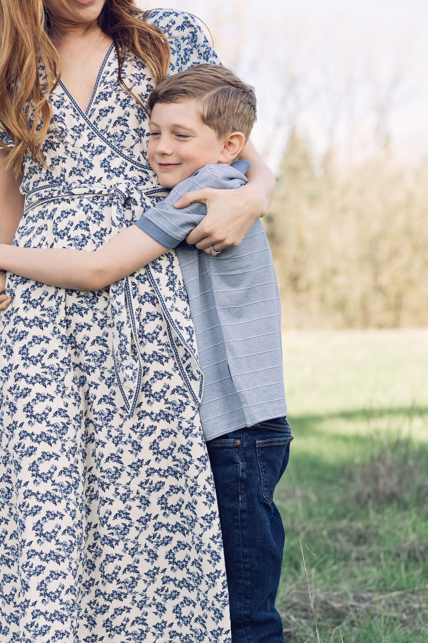 Young boy smiling while hugging his mothers waist in a bright outdoor spring setting.