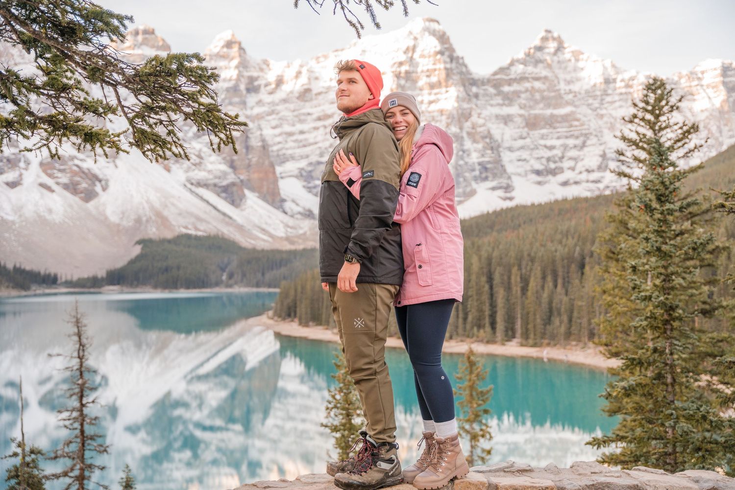 Couple standing on rocks at Moraine Lake, Banff, with turquoise water and snow-capped mountains in background.