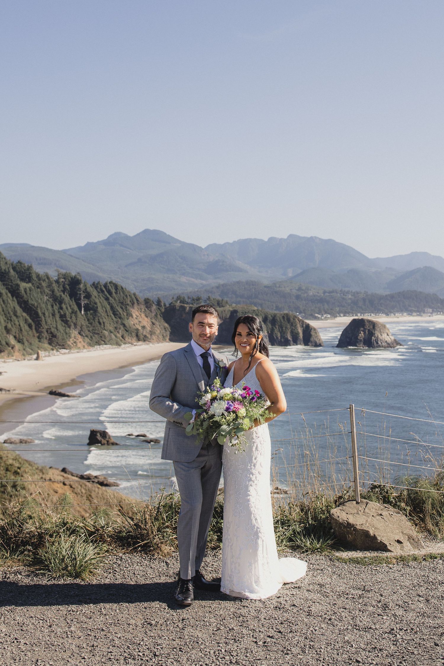 Bride and groom posing with bouquet on Oregon coast overlook with beach, sea stacks, and mountains backdrop.