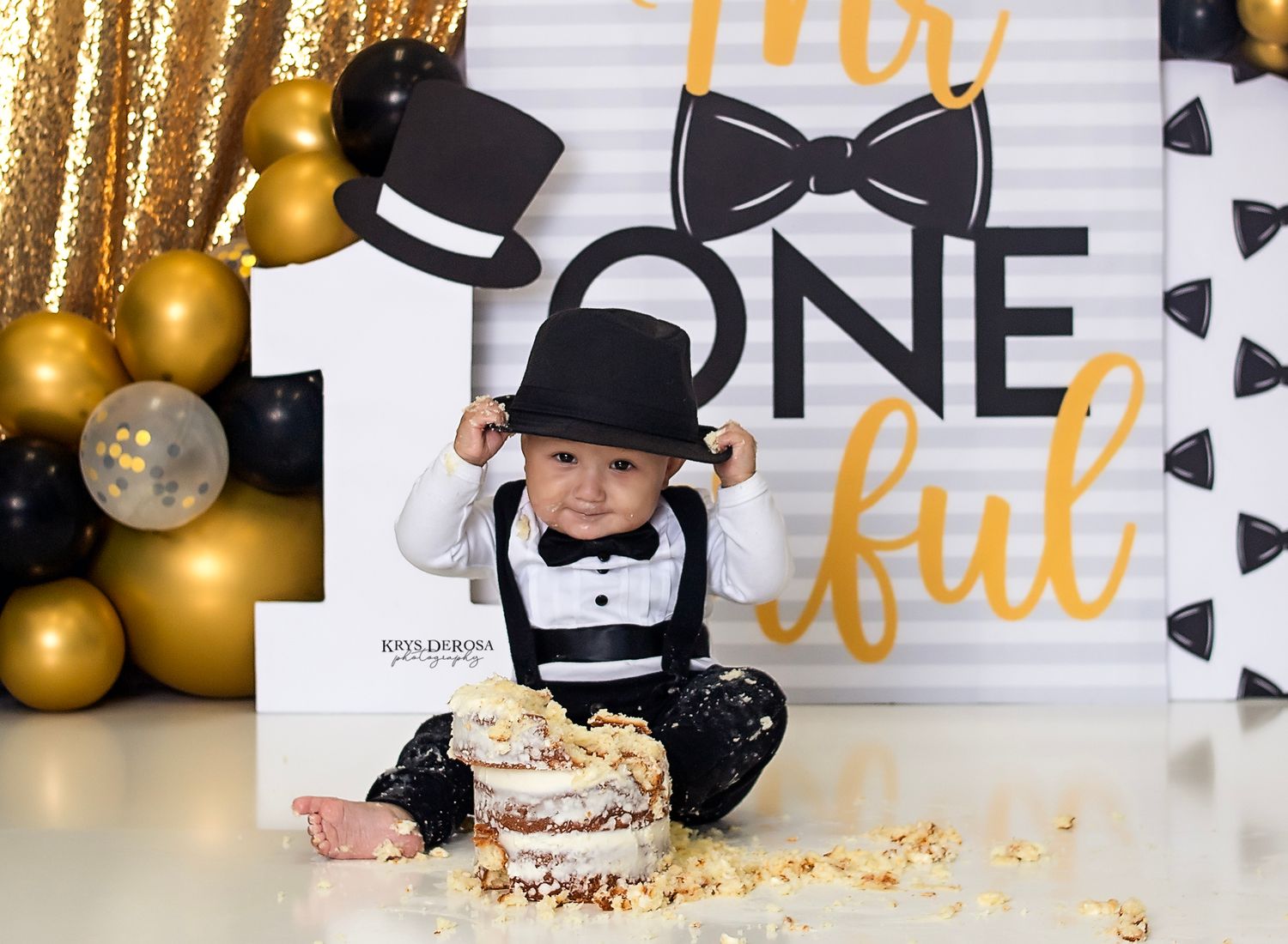 Baby boy in tuxedo and top hat sits beside smashed first birthday cake in front of Mr. ONEderful backdrop.