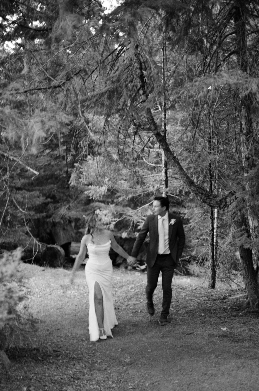 Black and white photo of a bride and groom holding hands walking along a forest path on their wedding day.