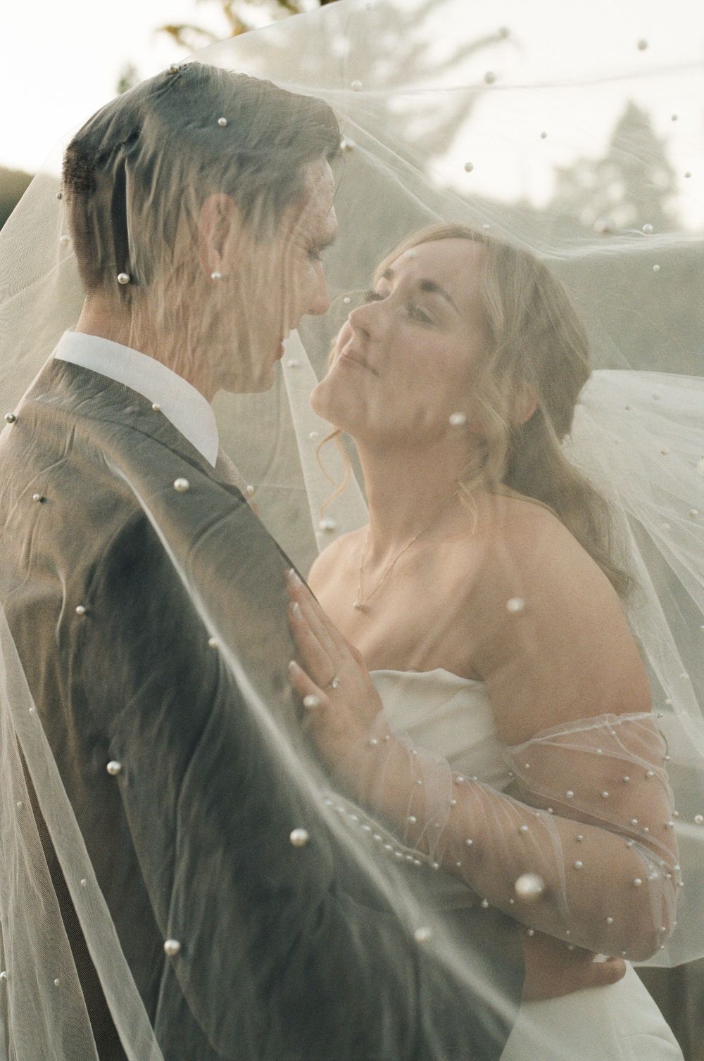Bride and groom share a romantic moment under a pearl-adorned wedding veil outdoors.