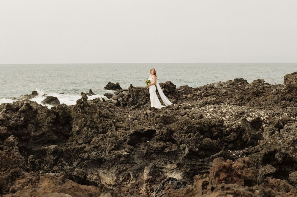 Bride in white wedding dress standing on rugged volcanic rocks by the ocean on a cloudy day.