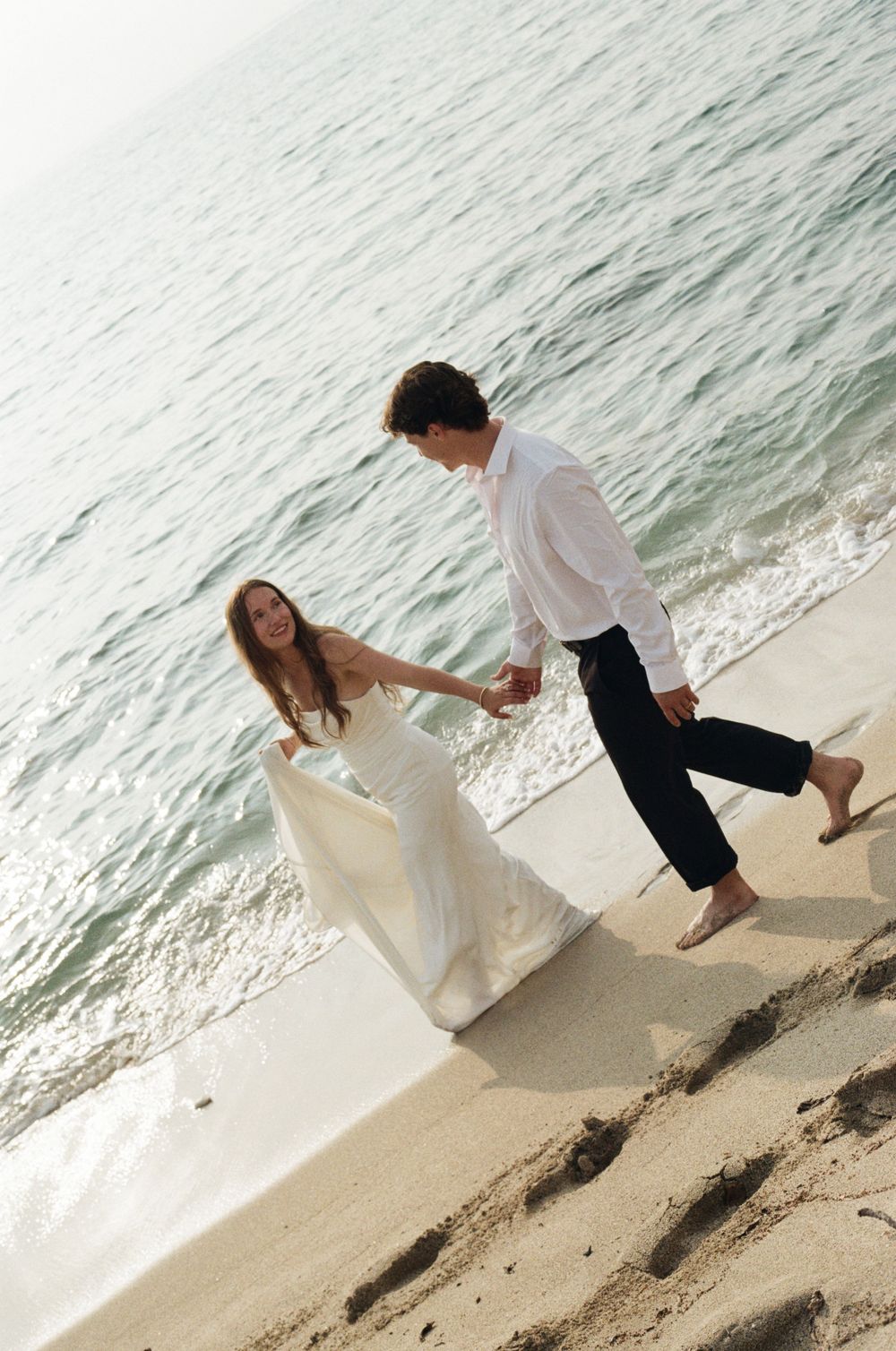 Newlywed couple walking hand in hand on a sandy beach, bride in white gown, groom in white shirt and dark pants.