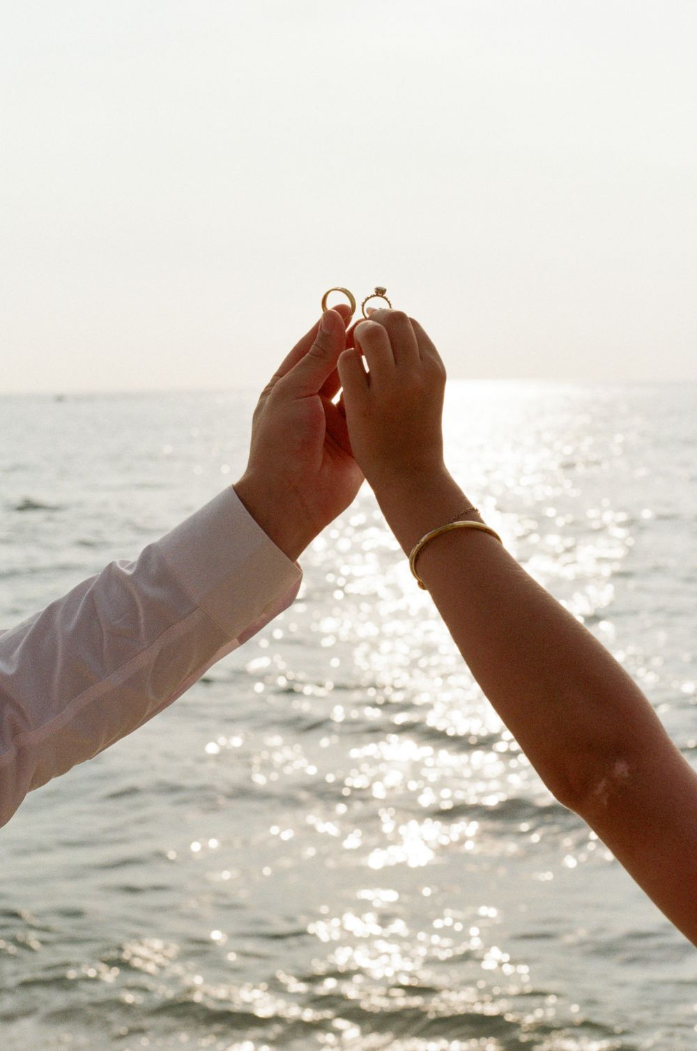 Couple holding wedding rings together over sparkling ocean water at sunset, romantic beach proposal scene.