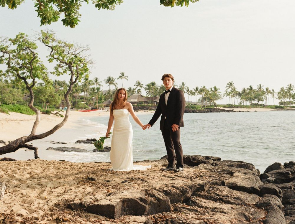 A bride and groom hold hands on rocky shoreline with tropical beach, palm trees, and ocean in background.