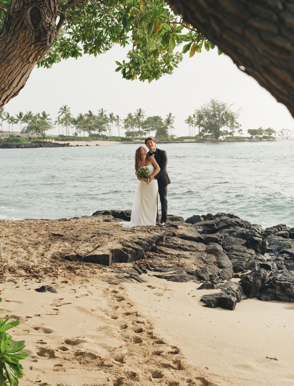 Bride and groom share a romantic kiss on a sandy Hawaiian beach, framed by lush trees and volcanic rocks.