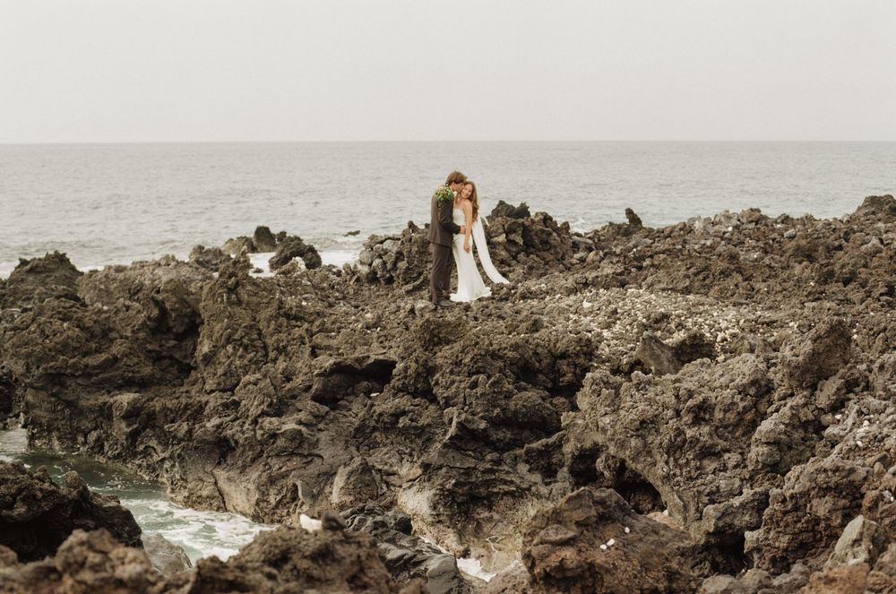 Couple in wedding attire embracing on rocky coastal shoreline with ocean waves and overcast sky backdrop.
