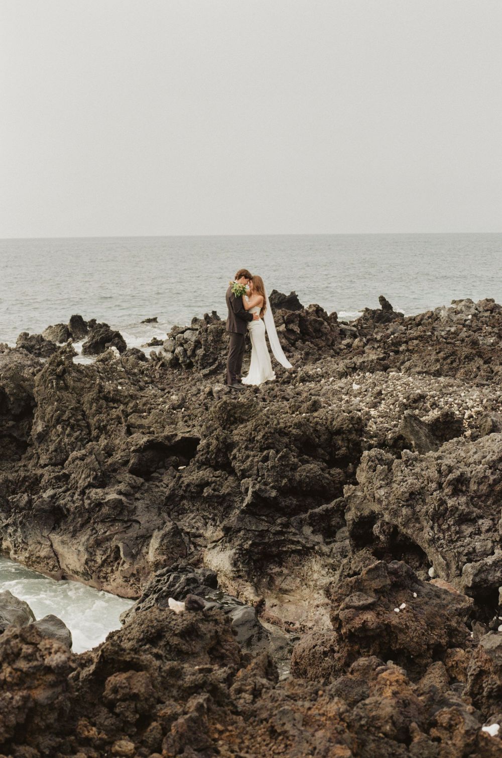 A bride and groom embrace on rugged volcanic rocks by the ocean during their romantic coastal elopement ceremony.