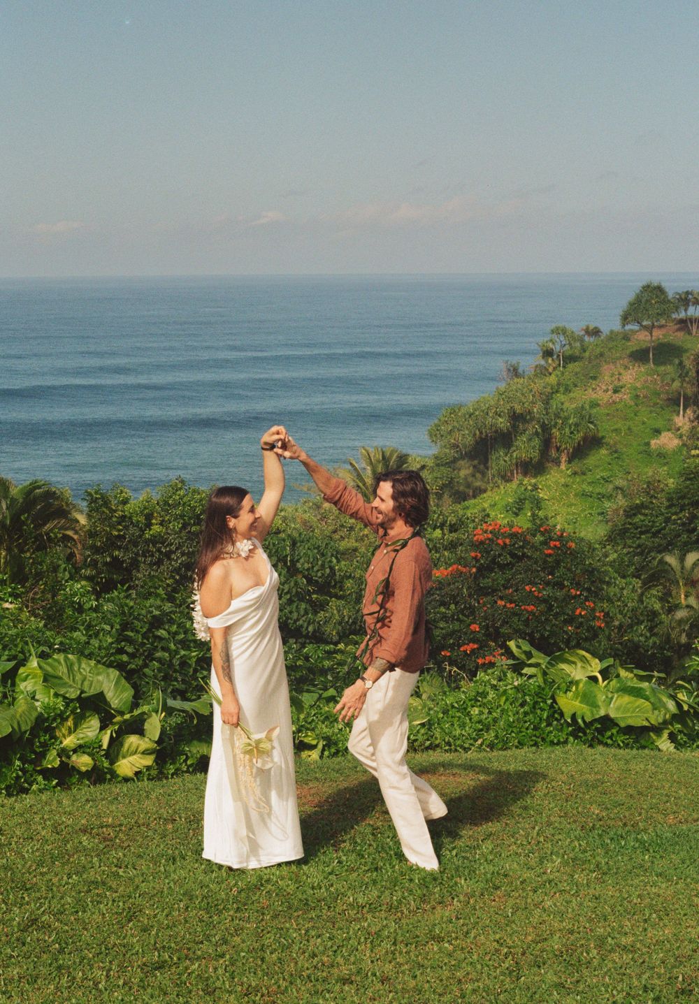 Couple dancing on a lush green cliff overlooking the ocean, bride in white dress holding flowers.