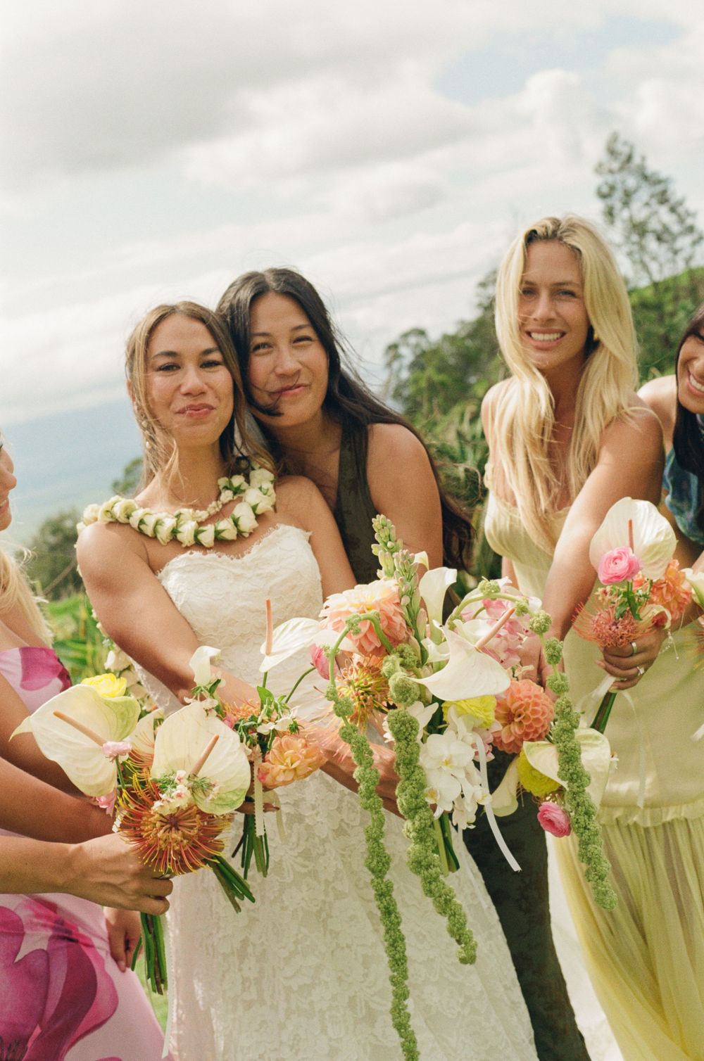 Bride in white lace gown with floral lei poses with bridesmaids holding tropical bouquets at outdoor wedding.