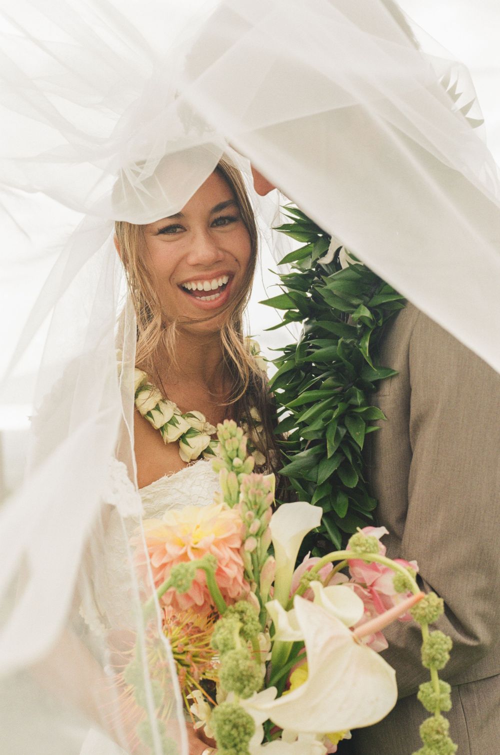 Smiling bride wearing a floral lei holds a pastel bouquet under a white veil at a tropical wedding ceremony.