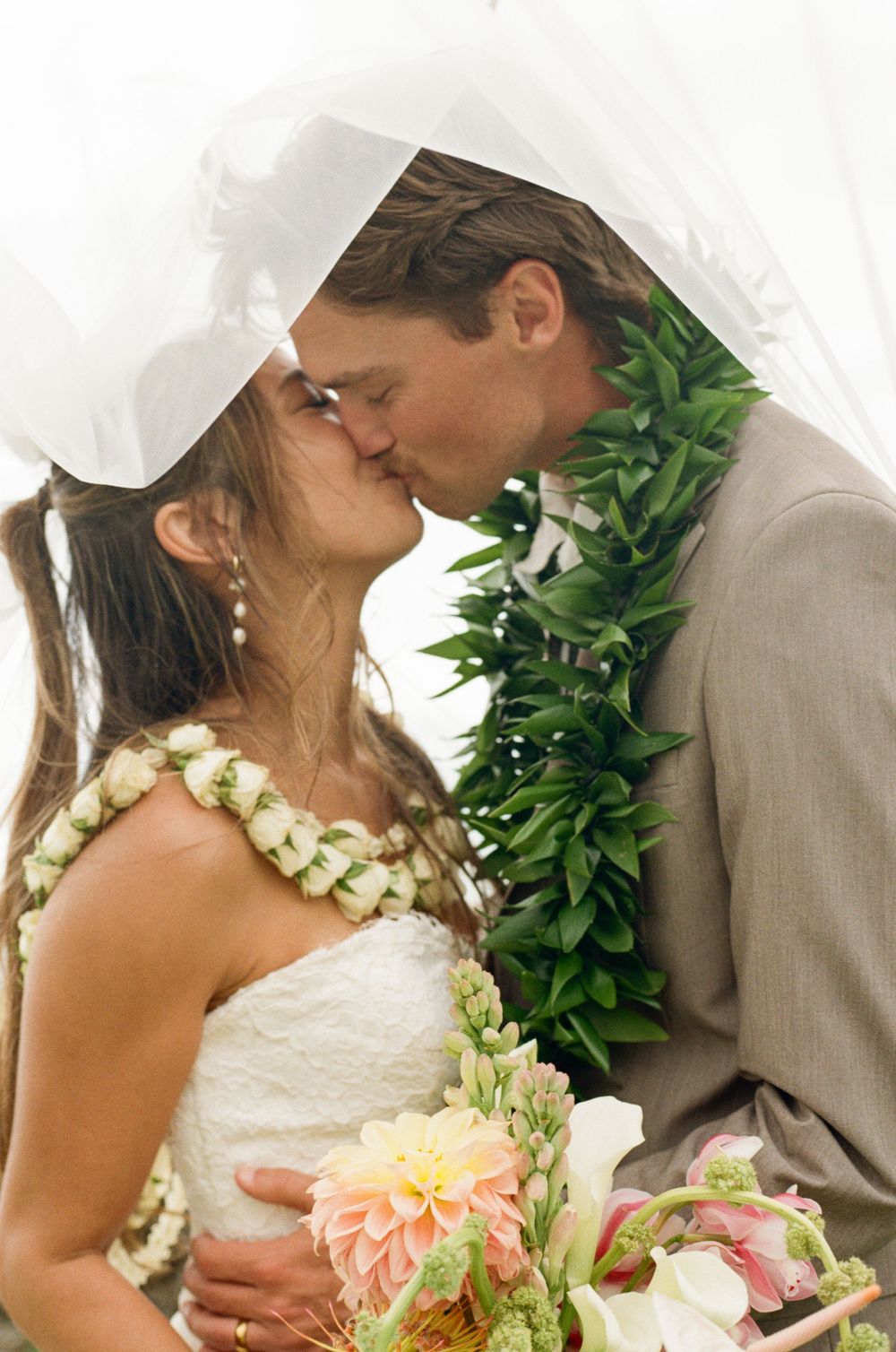 Bride and groom sharing a kiss under a white veil, wearing floral leis, with the bride holding a pastel bouquet.