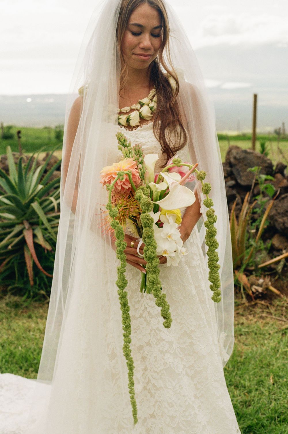Bride in lace wedding dress and veil holds tropical bouquet with green amaranthus and calla lilies outdoors.