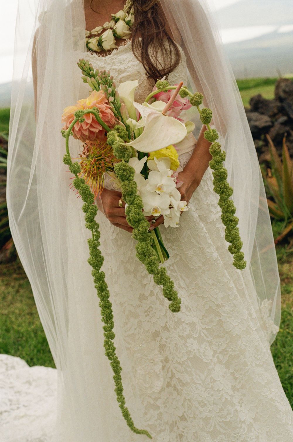 Bride in lace wedding gown holding tropical bouquet with calla lilies, pink flowers, and cascading green amaranthus.