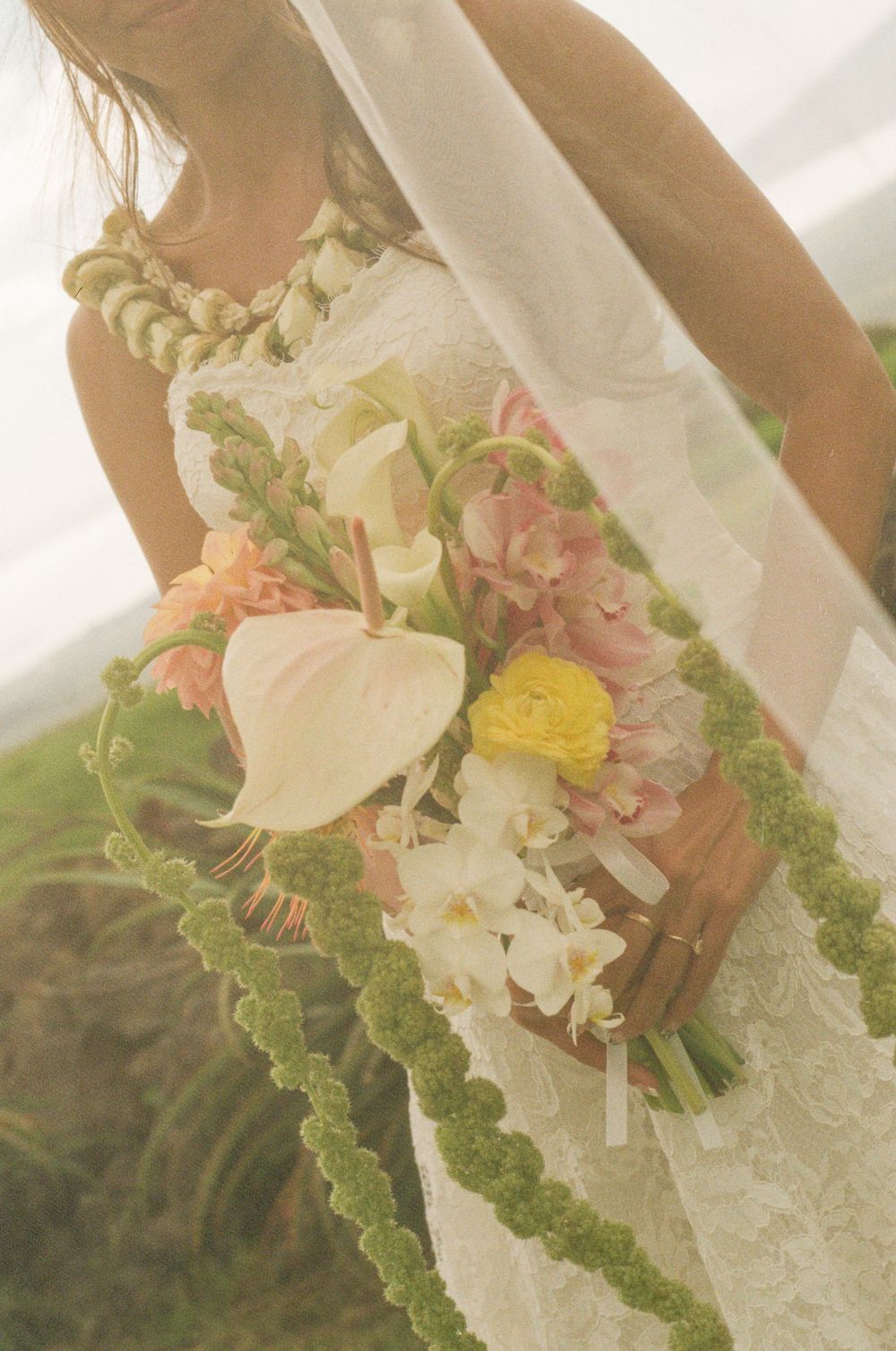 Bride holding tropical wedding bouquet with calla lilies, ranunculus, and orchids, adorned with green lei necklace.