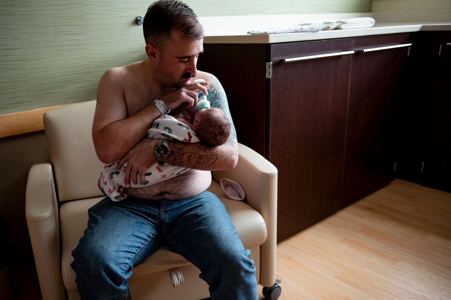 A parent wearing jeans holds and feeds a newborn baby while sitting in a hospital chair.