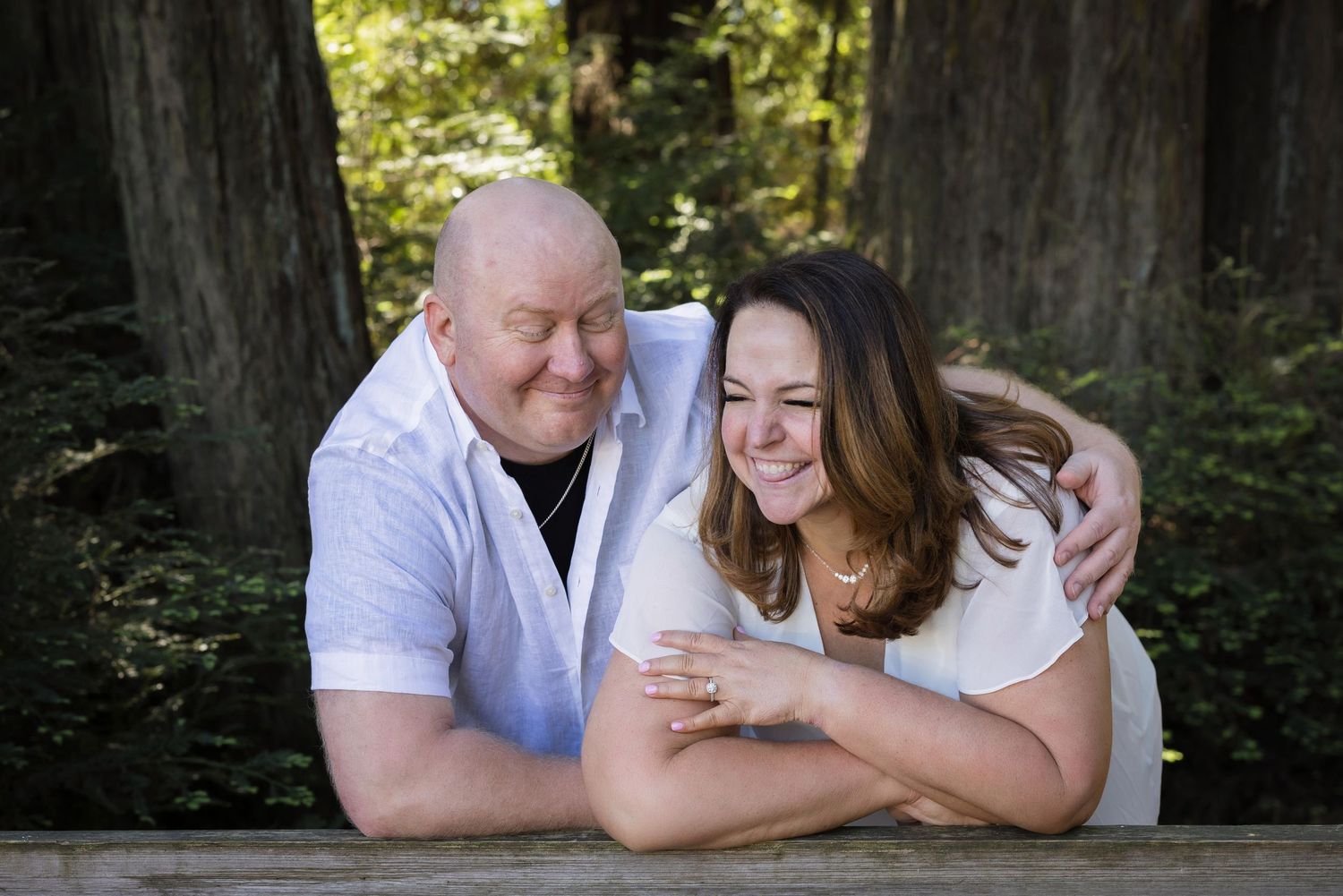 A smiling couple leans on a wooden railing outdoors, surrounded by tall trees, sharing a joyful moment together.