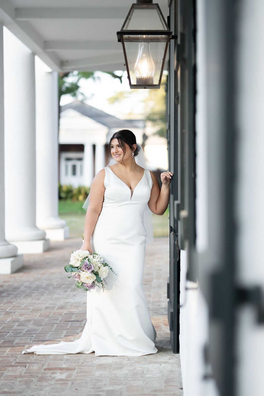 Bride in white sleeveless gown holding bouquet of roses, posing under lantern on elegant columned porch.