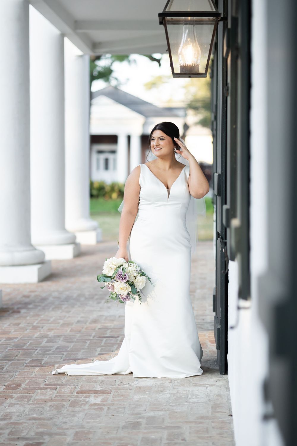 Bride in white sleeveless gown holding floral bouquet stands by column and lantern on brick walkway.