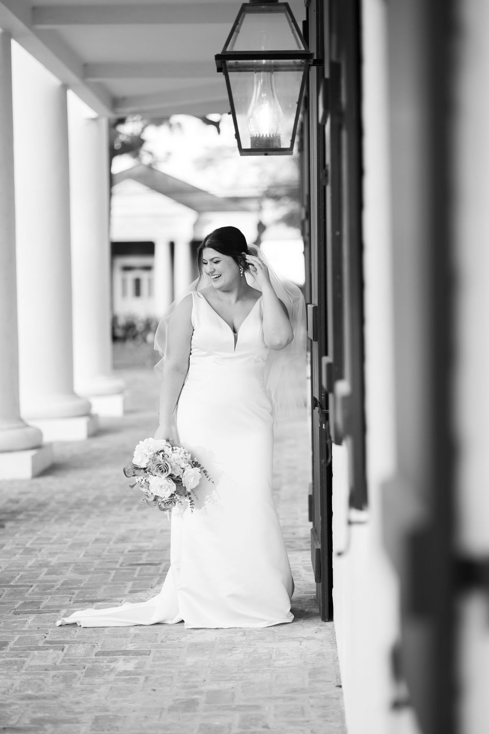 Black and white photo of bride in elegant white gown holding bouquet beside lantern and columns.